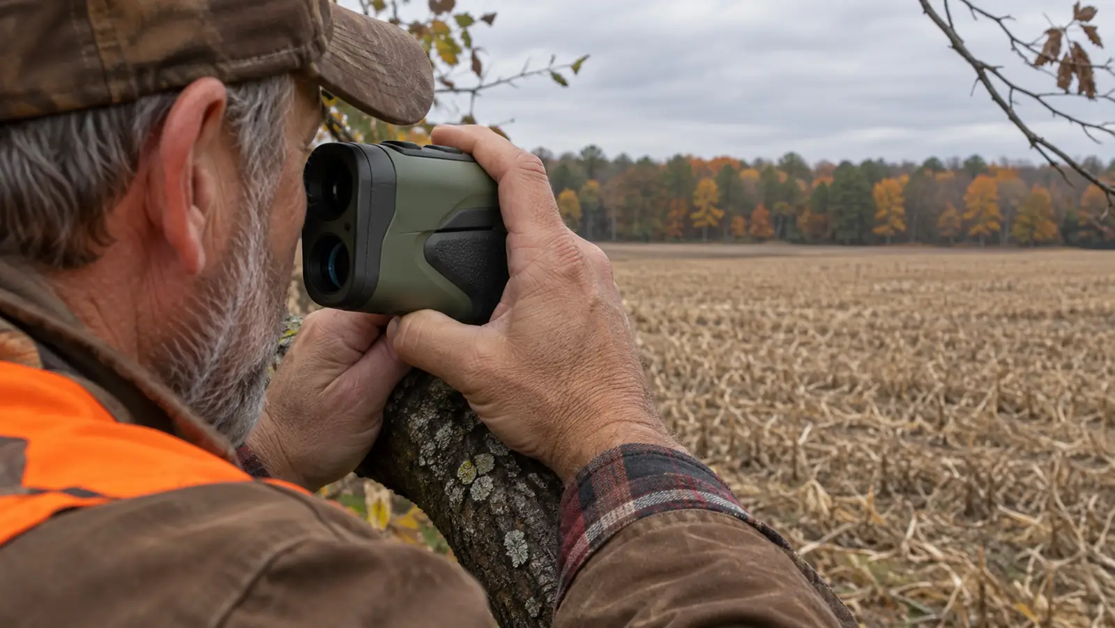 A hunter's hands holding a camo rangefinder steady against a tree branch while ranging a field edge