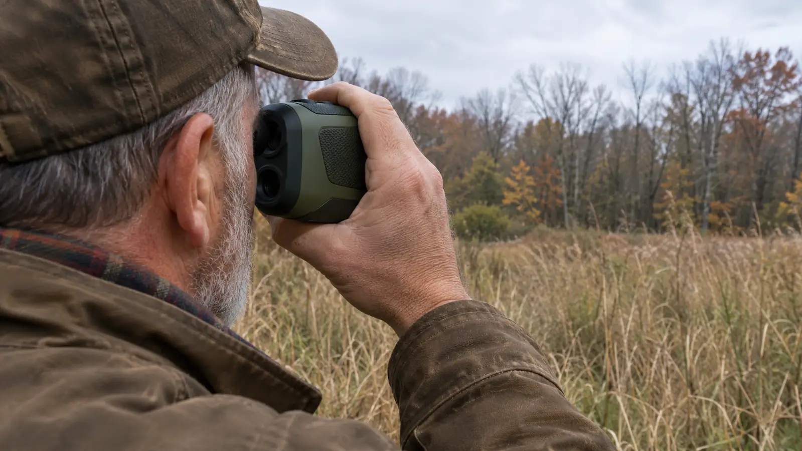 A hand holding a rangefinder up to one eye with tall grass and a field edge in the background